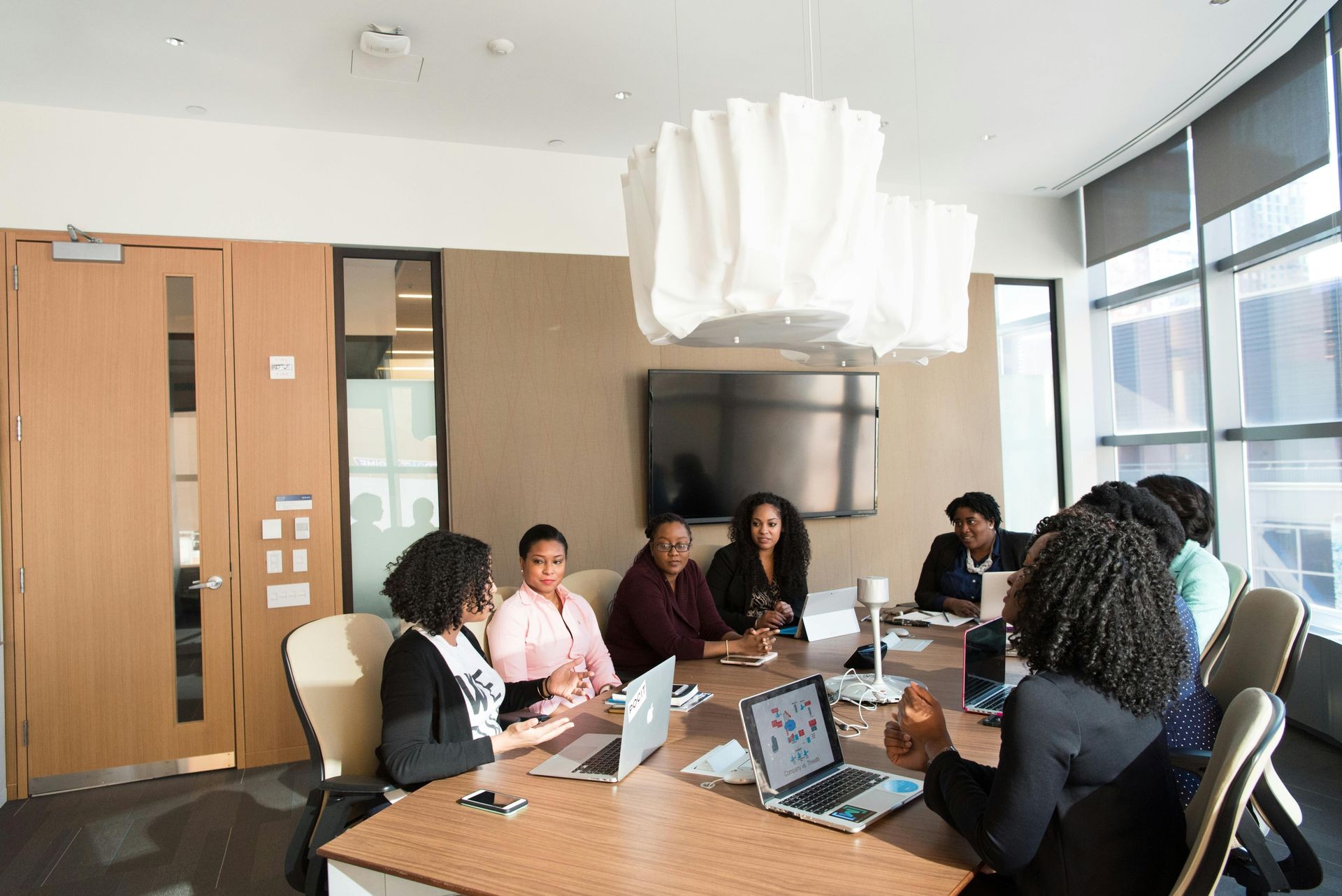 A group of Black women sit around a large conference table in a modern office meeting room. They are engaged in discussion, with laptops open in front of them. The room has floor-to-ceiling windows, a large flat-screen TV on the wall, and a unique white hanging light fixture above the table. The women appear to be participating in a professional meeting or collaborative session.